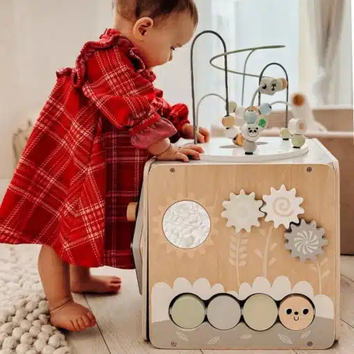 Montessori tegeluskuubik lastele 12+ kuud | E-pood Tootemaailm Beebitüdruk punases ruudulises kleidis seisab puidust tegevuskeskuse kõrval