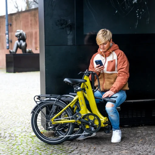 Inimene istub kollase elektriratta kõrval ja vaatab telefoni, taustal linnatänav