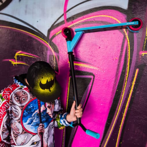 Trikitõukeratas Soke Go türkiissinine | E-pood Tootemaailm Laps hoiab trikiajalist tõukeratast värvilise grafiti seina ees