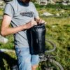 Lenksukott Giant H2Pro veekindel 12,5 l | E-pood Tootemaailm Mees pakib musta veekindlat kandekotti õues rohumaal, jalgratas taustal