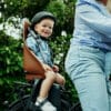 Laste jalgrattatool Urban Iki Bio pakiraamile | E-pood Tootemaailm Väike laps kiivris istub jalgratta lastetoolis täiskasvanu rattal õues