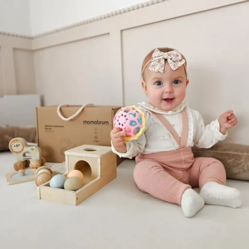 Montessori mänguasjakomplekt beebidele 7–12 kuud, 6-osaline | E-pood Tootemaailm Beebi istub mänguasjade keskel ja hoiab värvilist palli, taustal puidust mänguasjad ning Mamabrumi karp