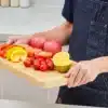Bambusest lõikelaud mahlasoone ja käepidemega 40 x 25 cm | E-pood Tootemaailm Põlluga inimene hoiab puidust lõikelauda viilutatud paprika, tomatite, õunte ja sidruniga
