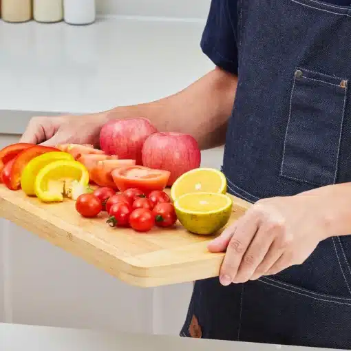 Bambusest lõikelaud mahlasoone ja käepidemega 40 x 25 cm | E-pood Tootemaailm Põlluga inimene hoiab puidust lõikelauda viilutatud paprika, tomatite, õunte ja sidruniga