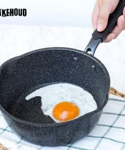 Foto mustast kastmepannist, milles on praetud muna; käsi hoiab panni sangast. Pann on must, täpilise mustriga ja asub heledal laual valge-rõhulise köögirätiku peal.