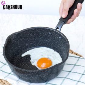 Foto mustast kastmepannist, milles on praetud muna; käsi hoiab panni sangast. Pann on must, täpilise mustriga ja asub heledal laual valge-rõhulise köögirätiku peal.